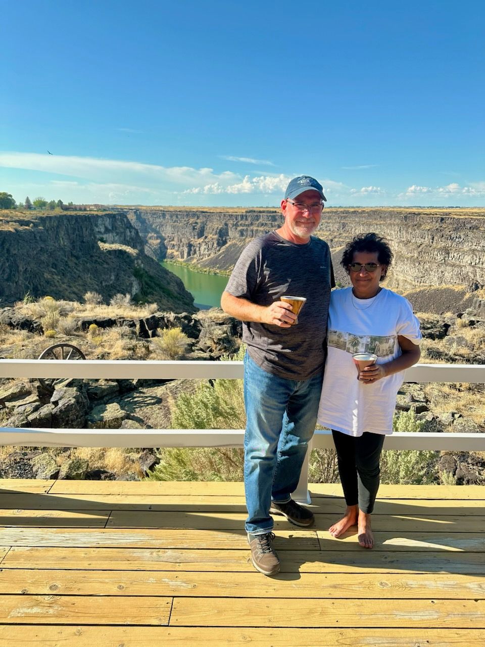 Couple on a wooden deck overlooking a canyon, holding drinks, under a blue sky.