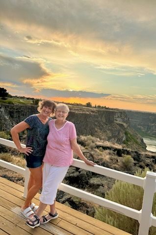 Two women pose with a sunset canyon view. One arm around each other; smiles. Wooden deck, white railing.