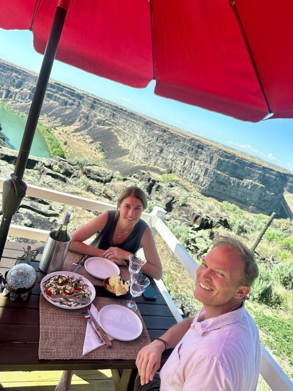 Couple dining outdoors under a red umbrella with a canyon view. Smiling, food on table.