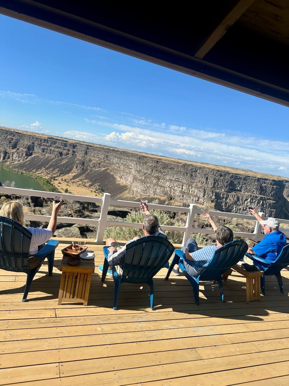 Four people relax on a deck overlooking a canyon. Blue chairs, sunny day, people waving.