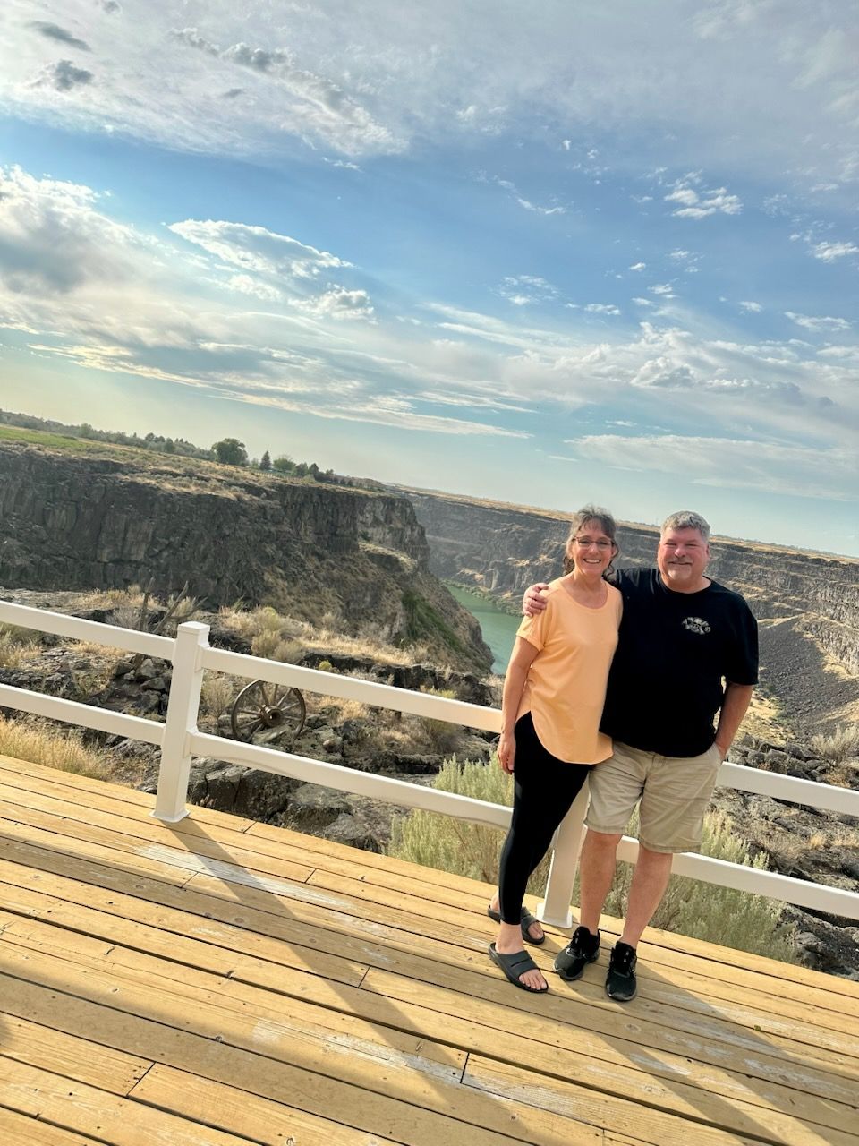 Couple on wooden deck overlooking a canyon with blue water under a cloudy sky.