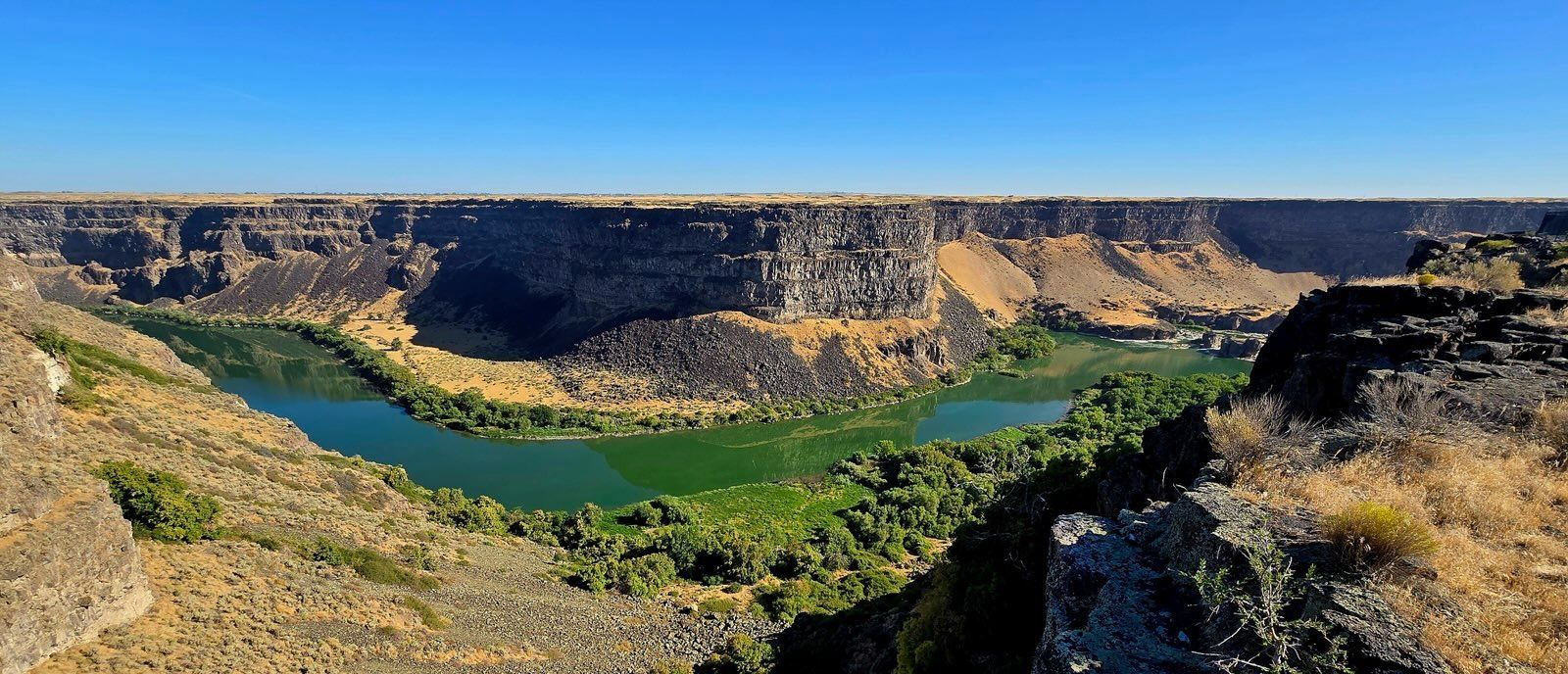 A wide canyon with a green river and blue sky. The canyon walls are brown and rocky.