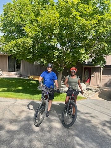 Two people on bikes in front of a house, under a large tree. One in blue, one in olive green.