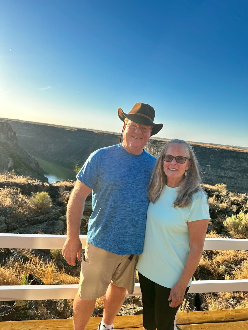 Two people smiling on a canyon overlook at sunset, with a blue sky and wooden railing.