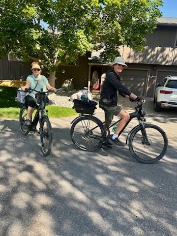 Two people riding black bicycles on a sunny street. They wear hats and have baskets. Houses in the background.
