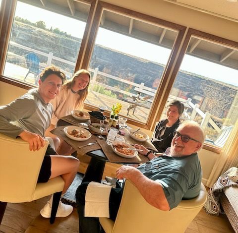Five people around a dining table by a sunny window, plates of food, relaxed smiles.