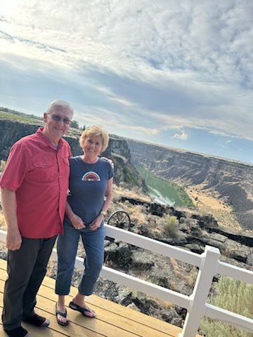Couple posing on a wooden deck overlooking a canyon. Man in red shirt, woman in blue, cloudy sky.