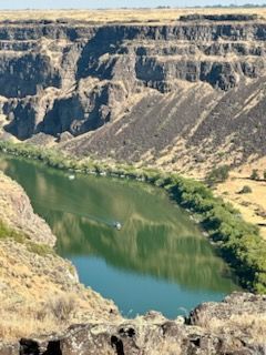 Canyon with green river and boats, under a blue sky. Rocky cliffs on both sides, dry brush.