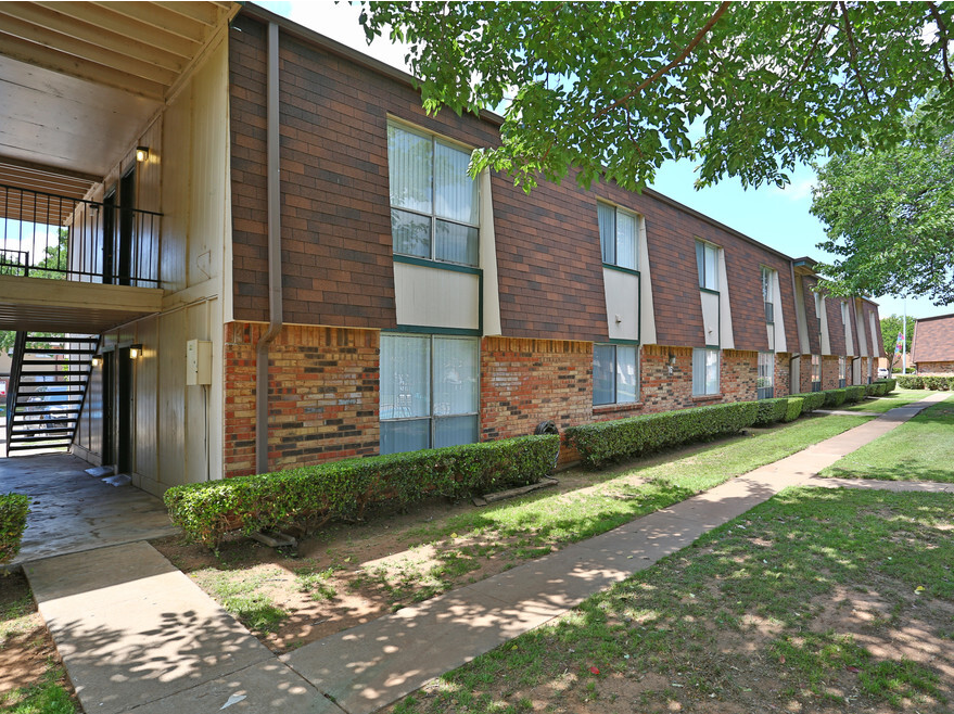 Apartment building with brick facade and green bushes lining a sidewalk.