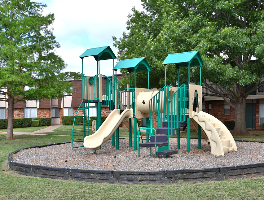 Playground with green and tan structures, surrounded by mulch, trees, and grass.