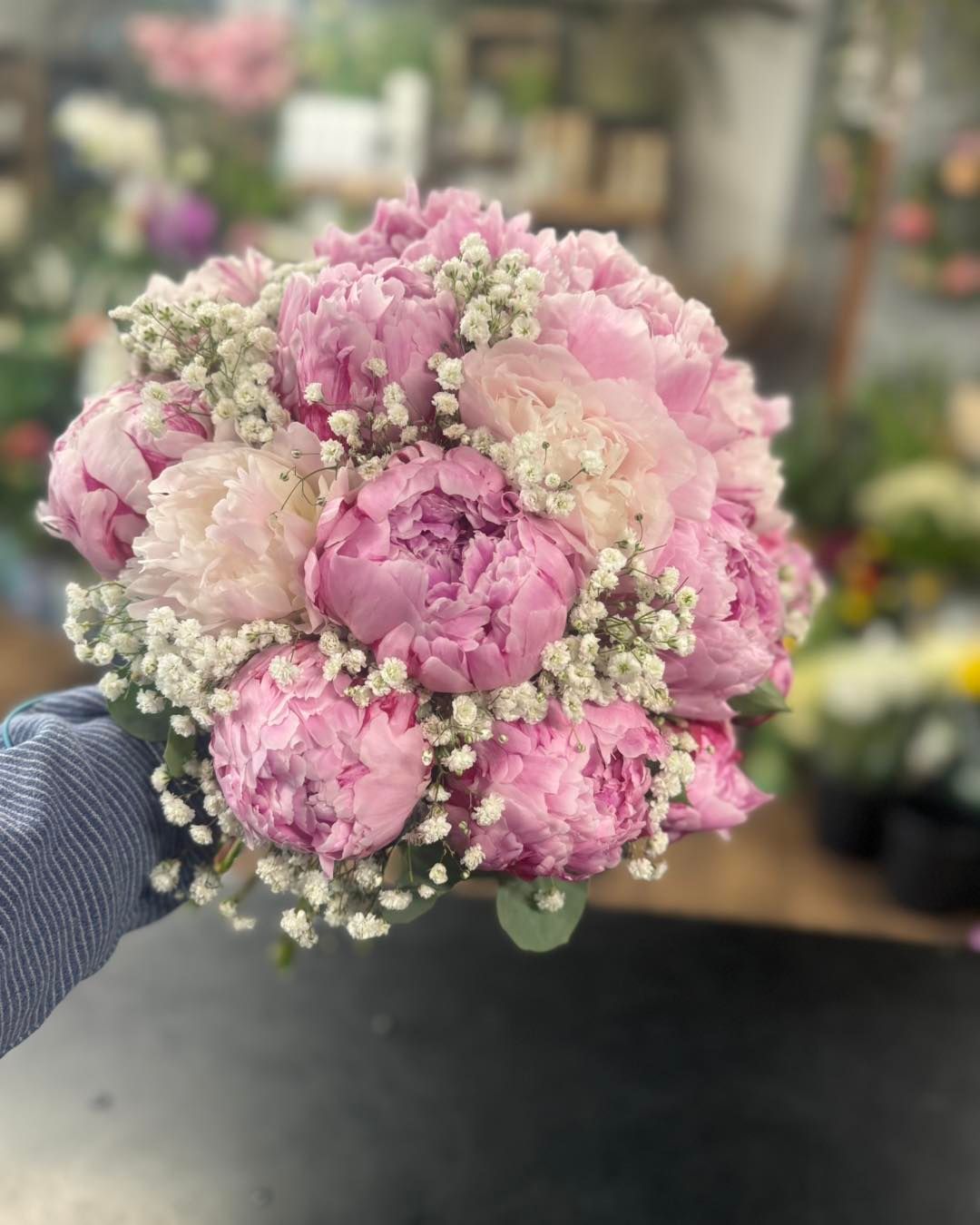 Pink peony bouquet with baby's breath, held against a blurred floral shop background.