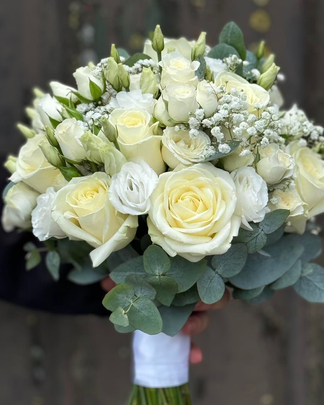 White rose and lisianthus bouquet wrapped with ribbon, held against a blurred background.