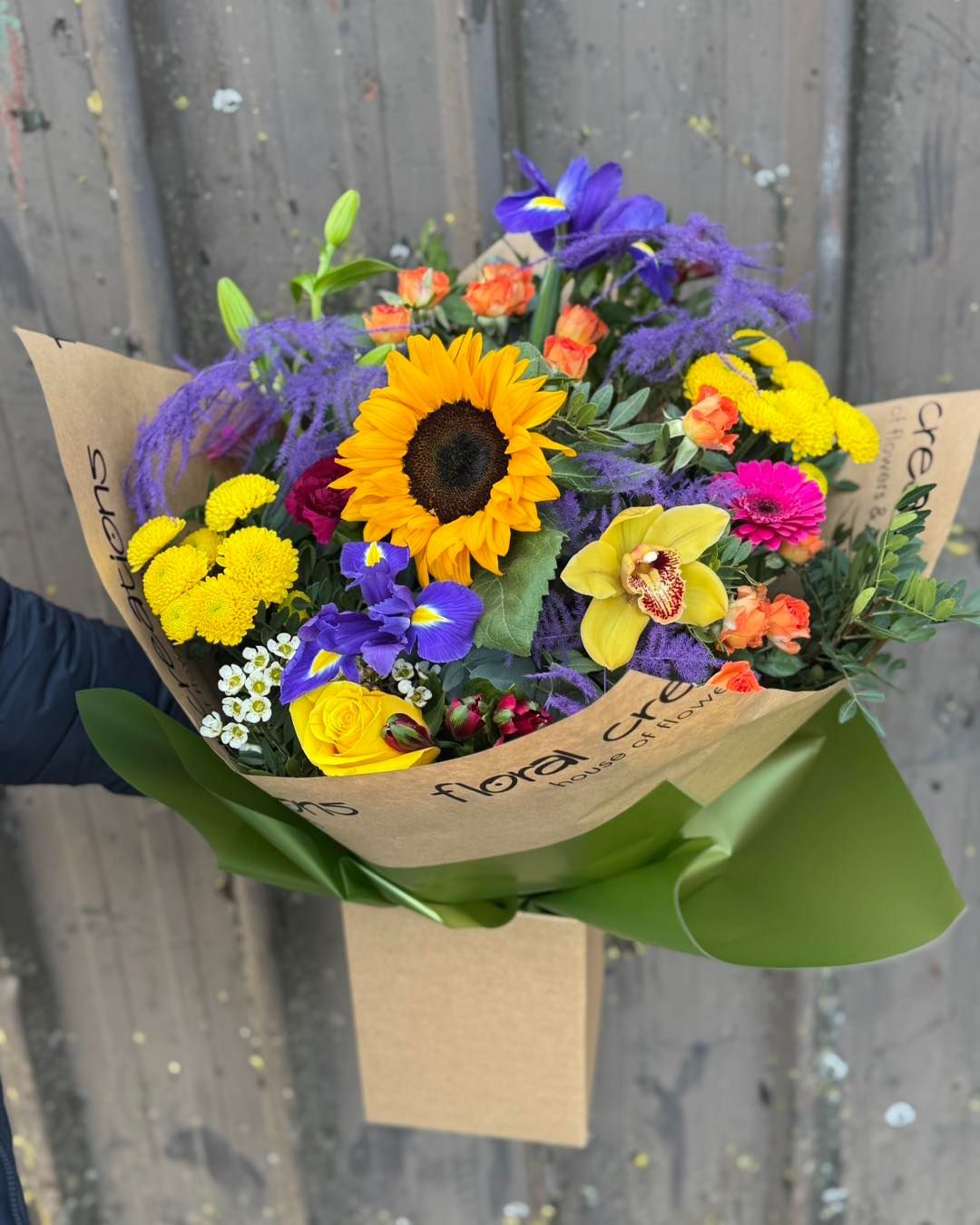 Colorful flower bouquet with a sunflower, irises, and other blooms, held in a brown paper wrap.