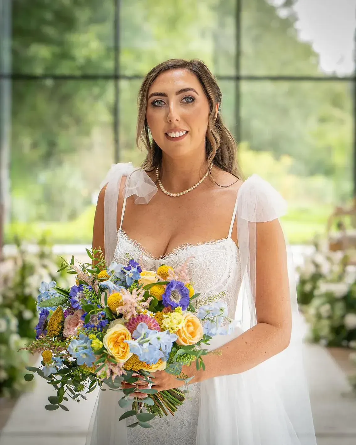 Bride holding colorful bouquet, wearing a white wedding dress with sheer sleeves, standing indoors.