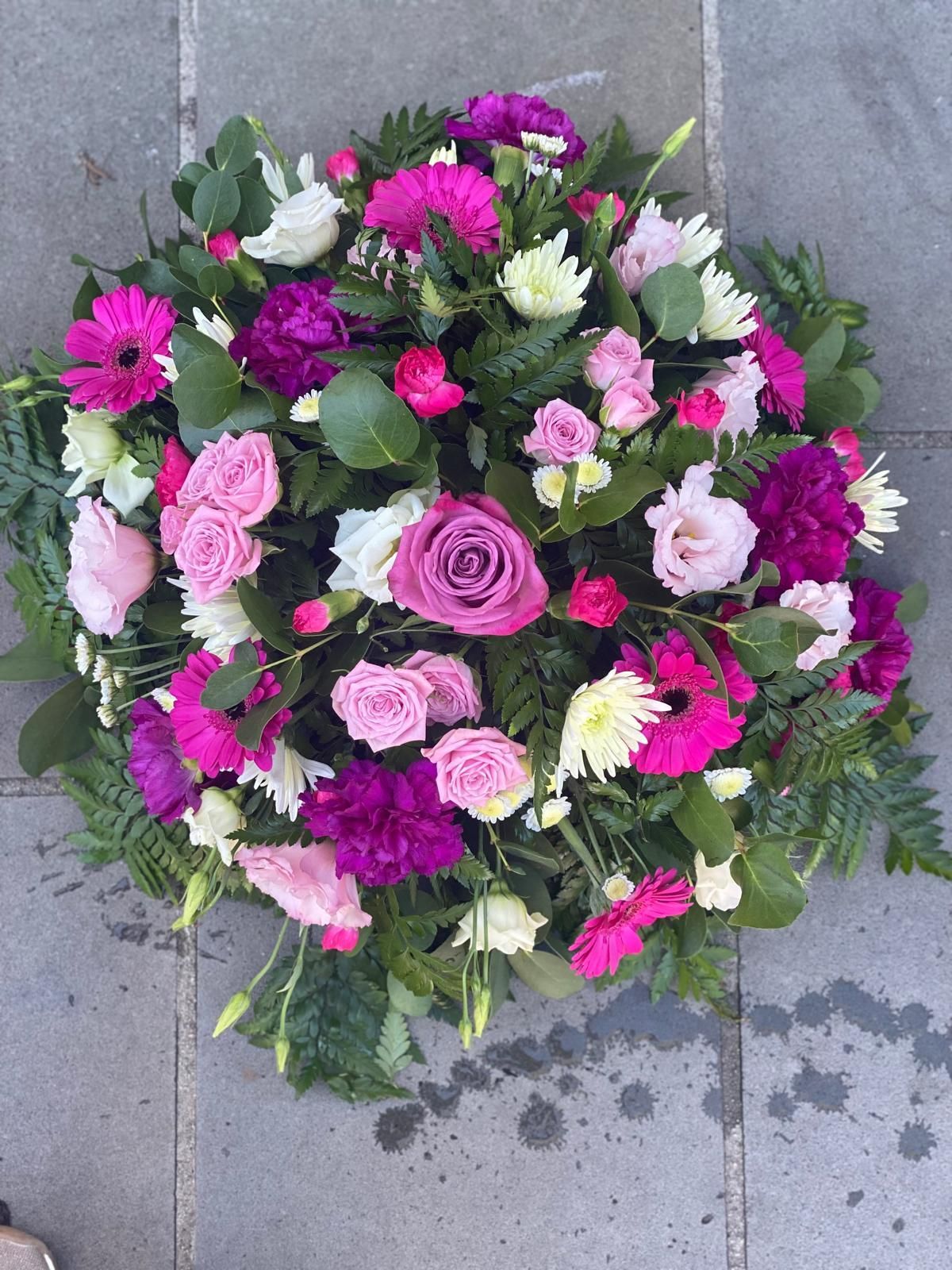 A floral wreath of pink, purple, and white flowers, including roses and gerberas, on a gray stone surface.