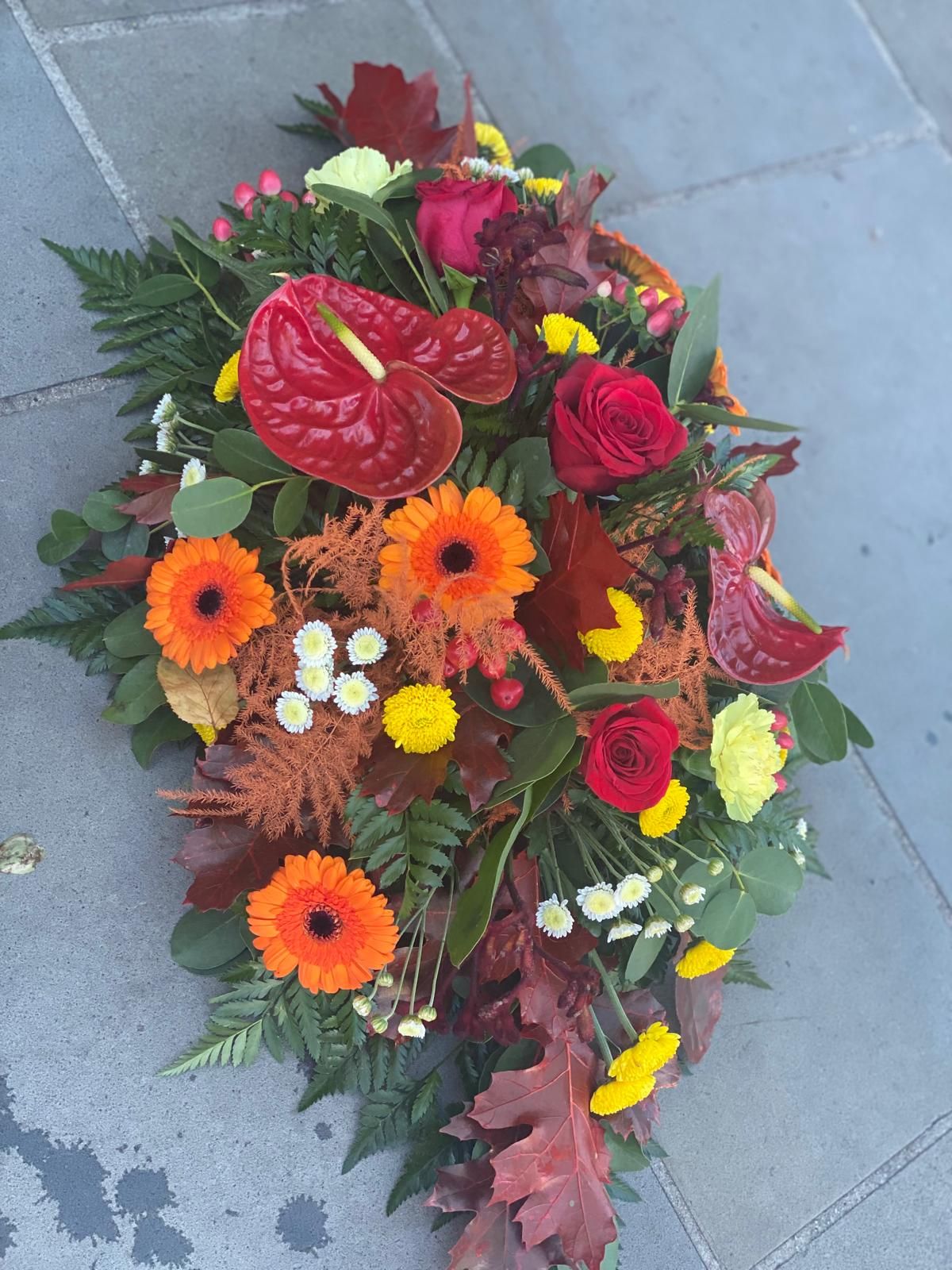 Floral arrangement with red, orange, and yellow flowers, surrounded by green foliage, resting on stone.
