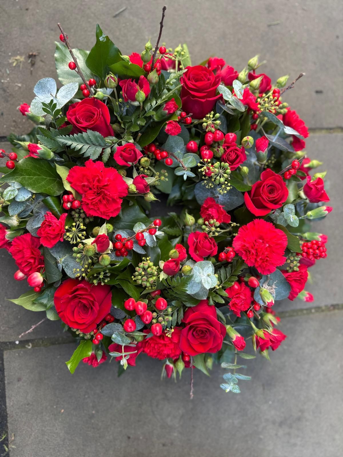 A circular floral wreath composed of red roses, carnations, berries, and greenery, on a stone surface.