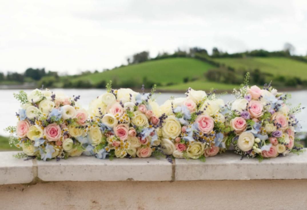 Four pastel-colored floral bouquets arranged on a white ledge overlooking water and a green hill.