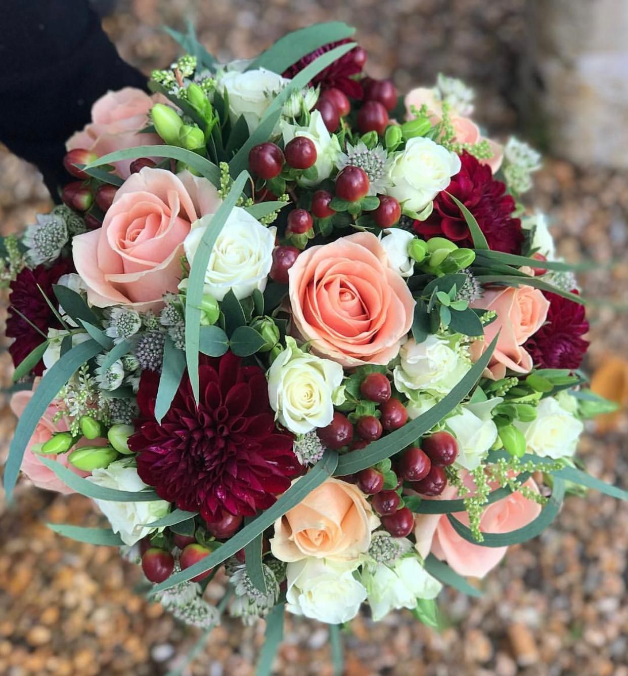 A round bouquet of peach, white, and burgundy flowers with green foliage and red berries.