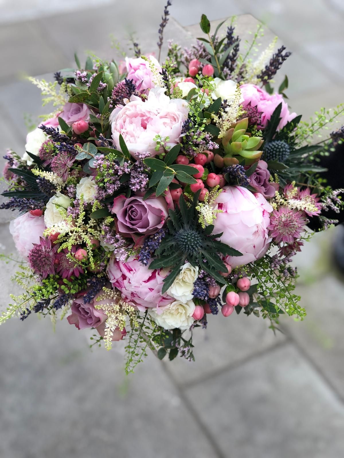 Bouquet with large pink peonies, purple roses, berries, and greenery, held against a grey background.
