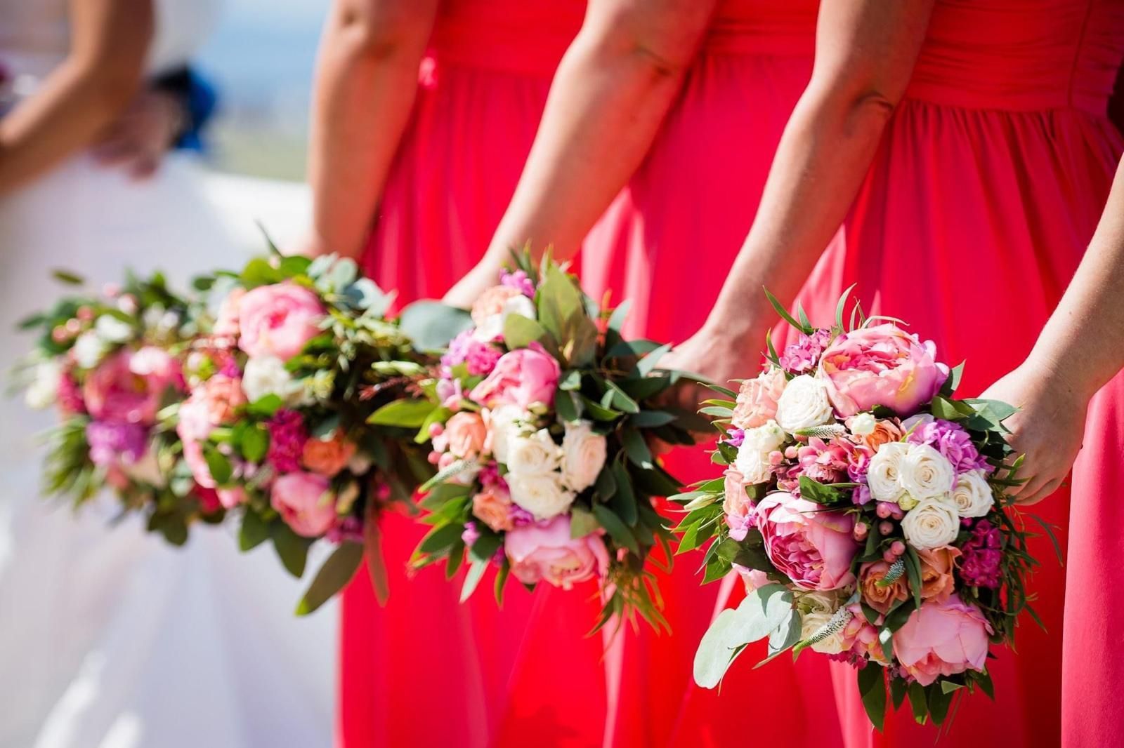 Bridesmaids holding bouquets of pink and white flowers, wearing coral-red dresses, in sunlight.