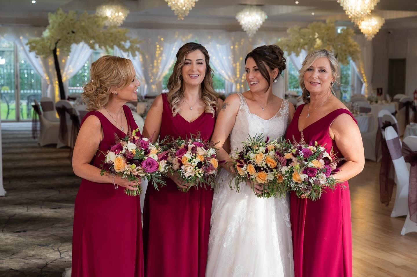 Bride and bridesmaids in burgundy dresses, holding bouquets, smiling at a wedding reception.