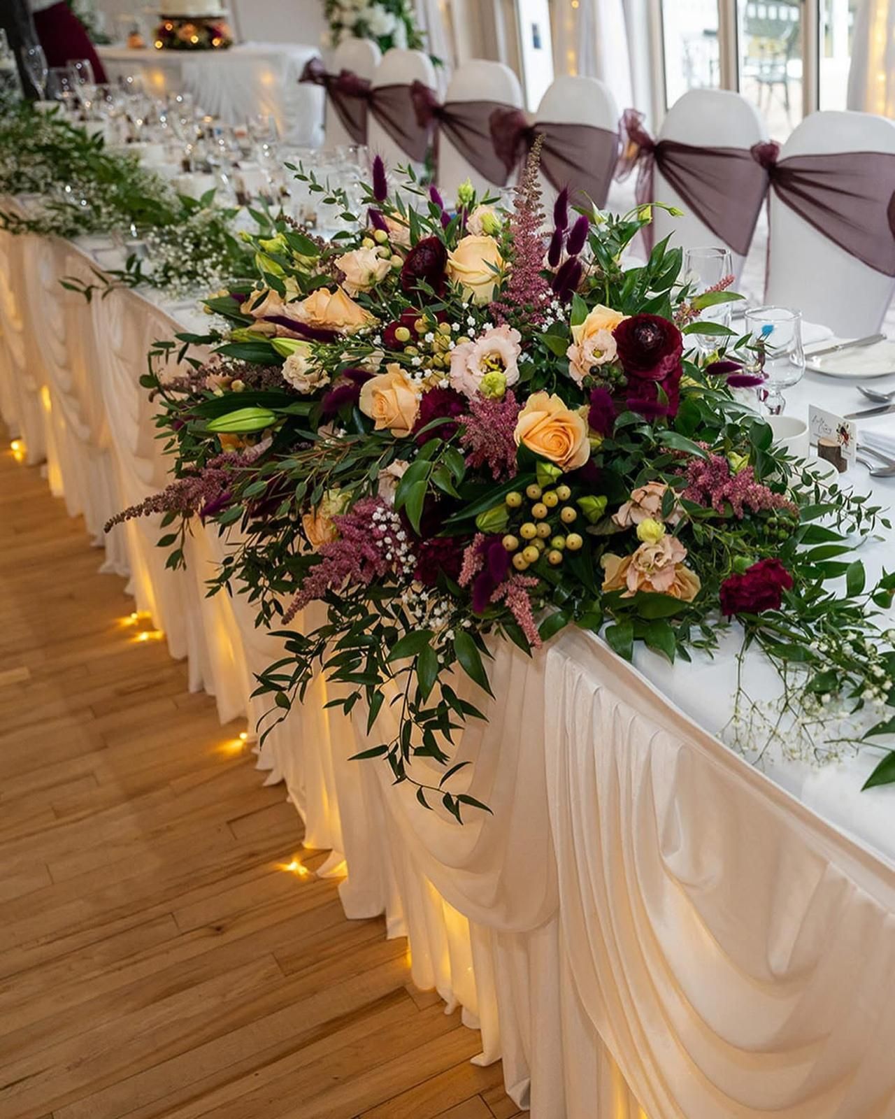 Wedding table decorated with flowers, white tablecloth, draped skirt, and burgundy chair sashes.