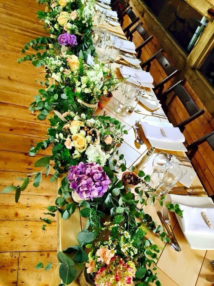 Long wooden table set for a formal event, decorated with floral arrangements and greenery.