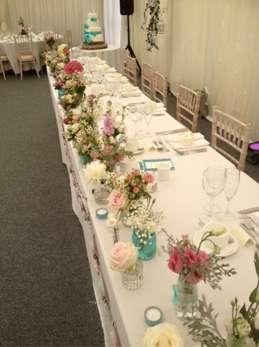 Long rectangular wedding reception table with floral centerpieces, cake, and place settings.