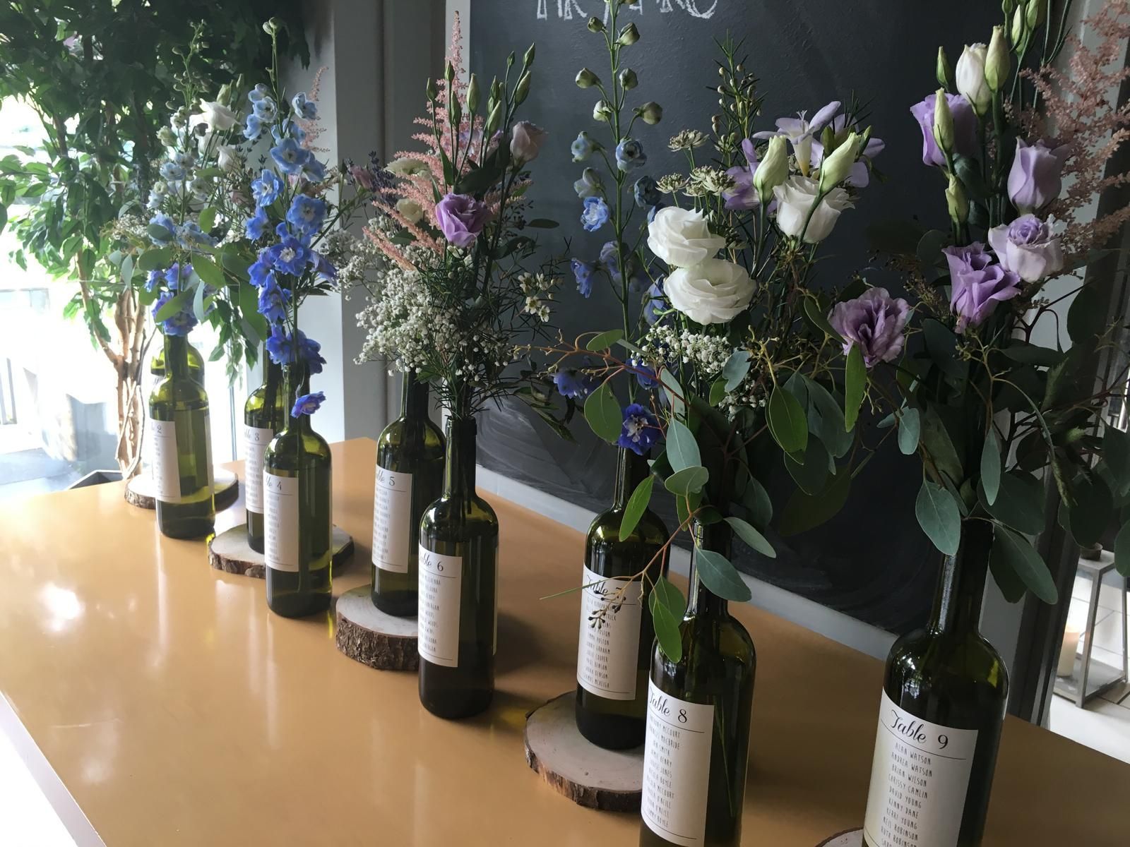 Floral arrangements in wine bottles on a wooden table, against a dark backdrop.