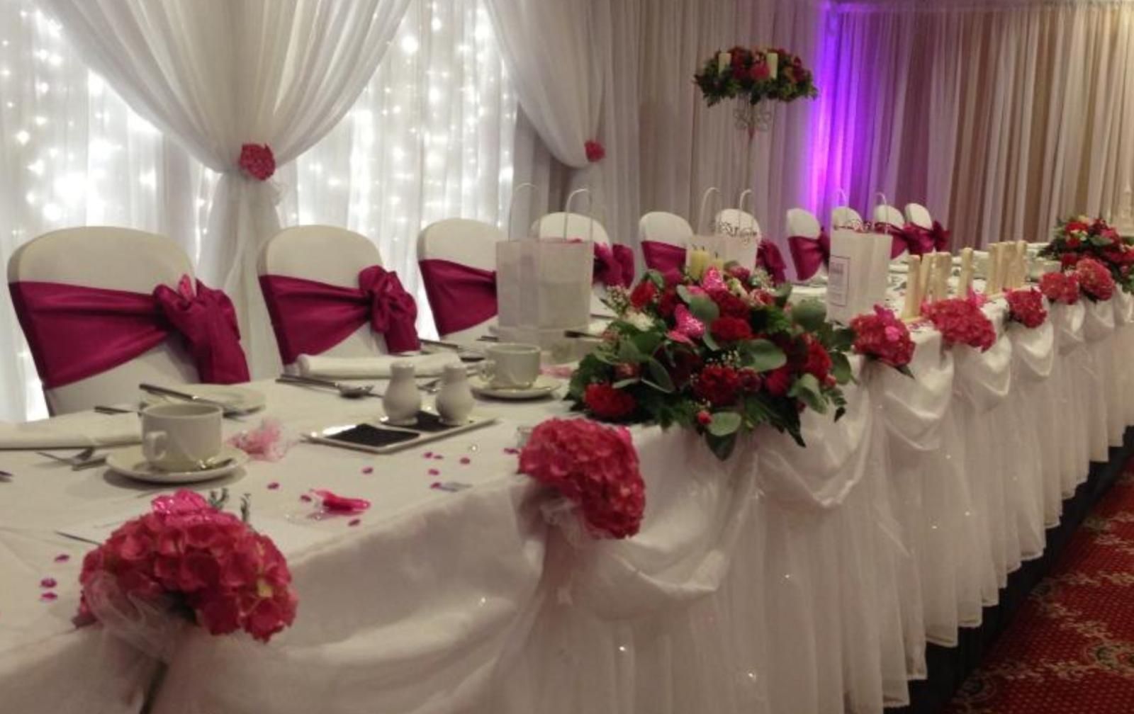 Wedding reception table, white tablecloth with magenta accents, flowers, and draped fabric.