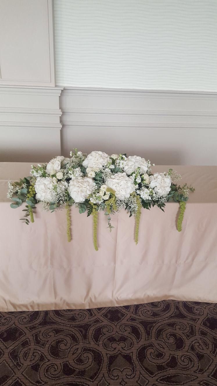 White floral arrangement on a beige-covered table with green accents; set against a light-colored wall.