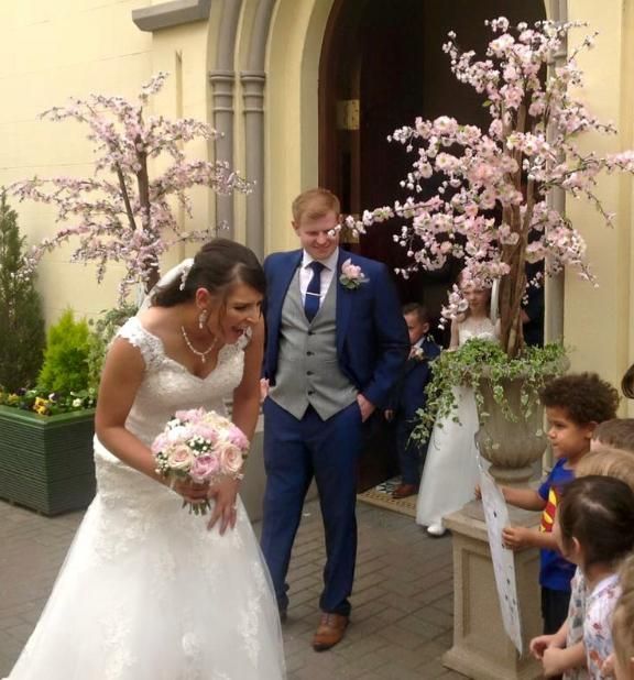 Bride and groom exiting a building, smiling. Flowers decorate the entrance. Children watch.