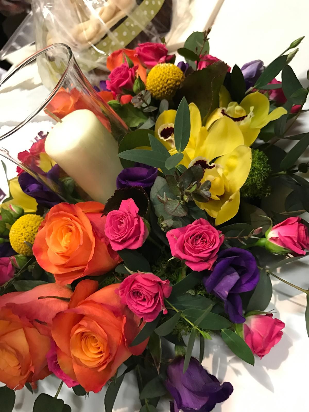 Vibrant floral arrangement with orange roses, yellow orchids, and a white candle.