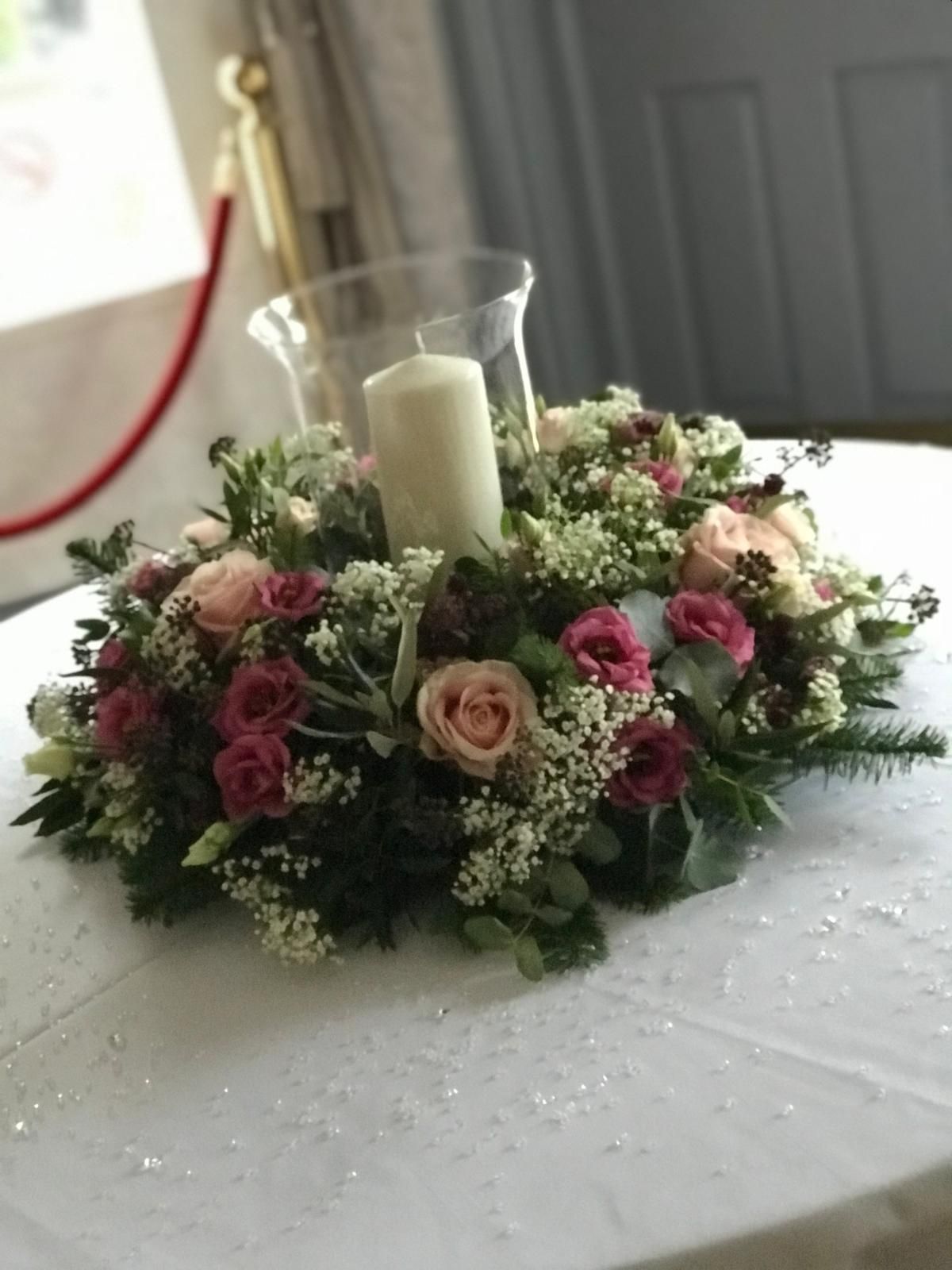 Floral centerpiece with candles on a white tablecloth; featuring pink and peach roses, greenery, and a tall glass hurricane.