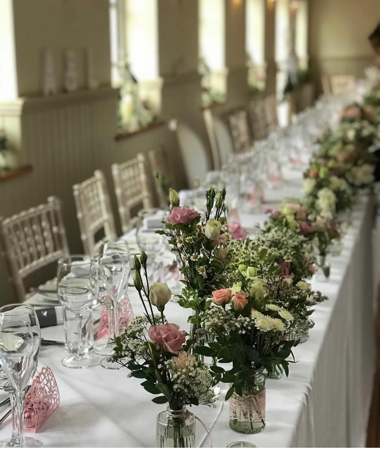 Long table set for a formal event, featuring flower arrangements, glassware, and white tablecloths in a bright room.