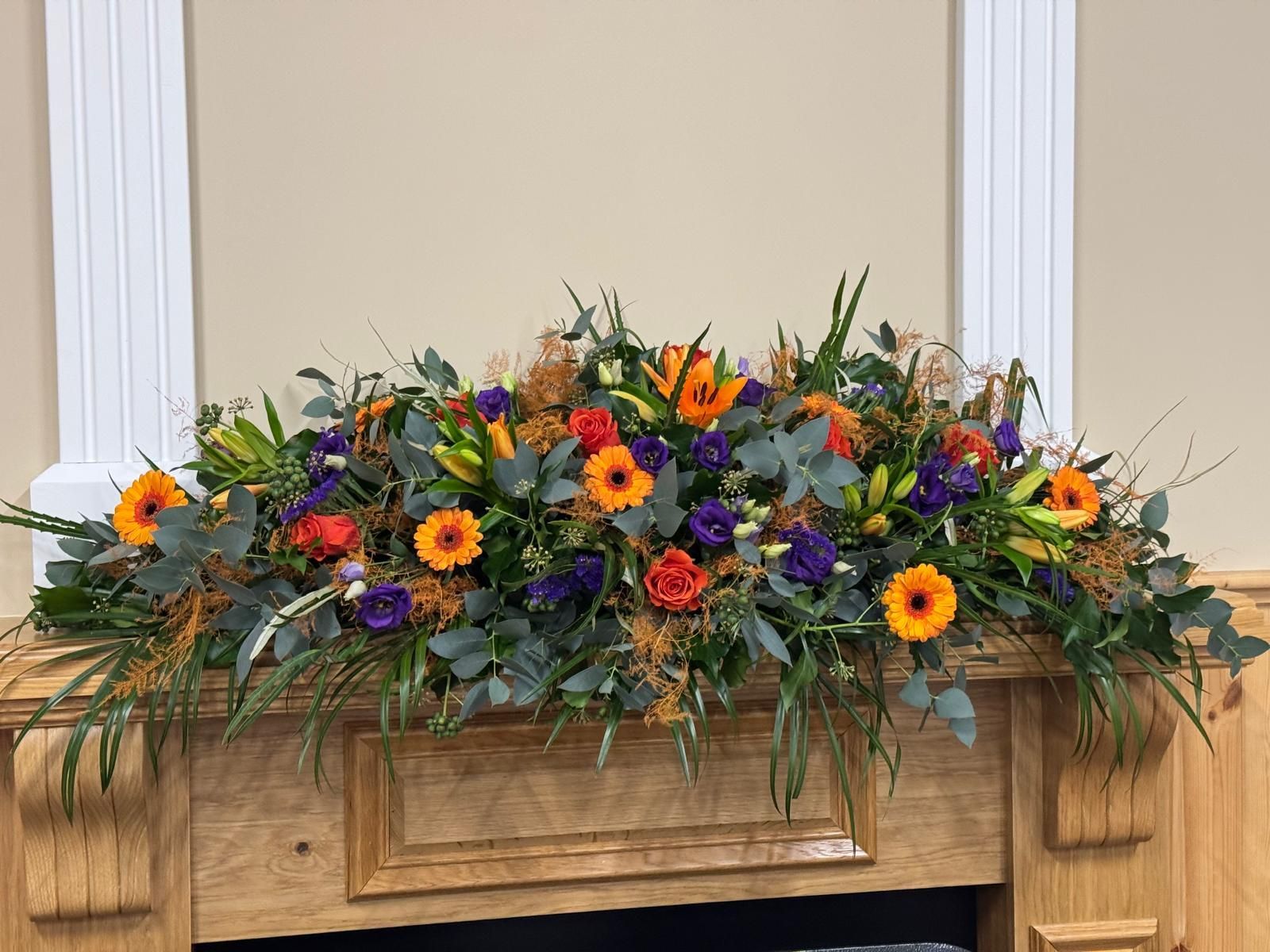 Floral arrangement of orange, purple, and green flowers on a wooden mantelpiece.