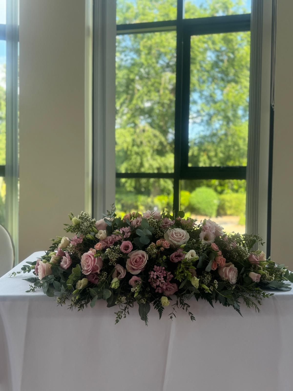 Floral arrangement of pink and green flowers on a white table, set before a large window.
