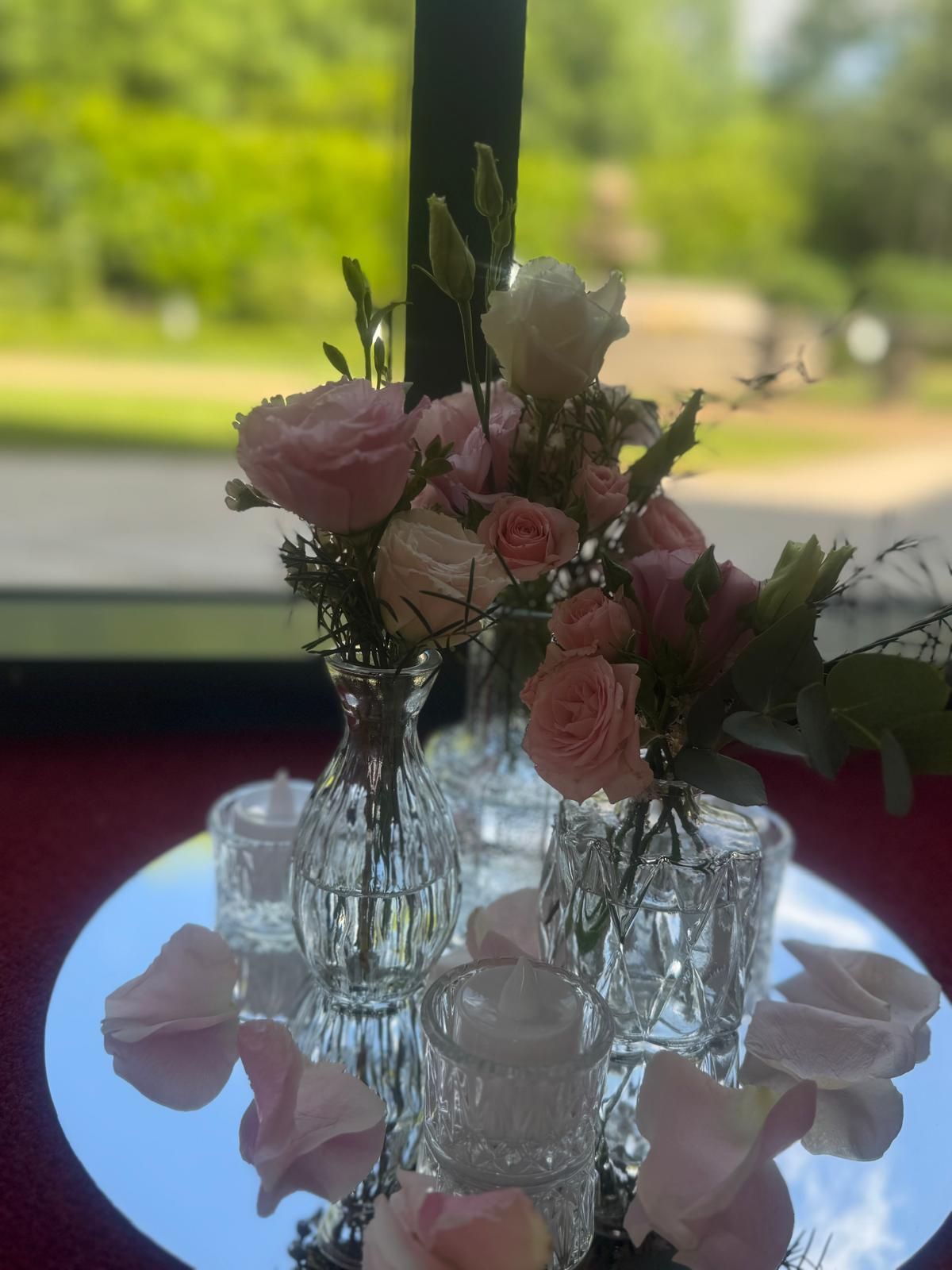 Pink and white flowers in glass vases on a mirrored surface with scattered petals and candles.