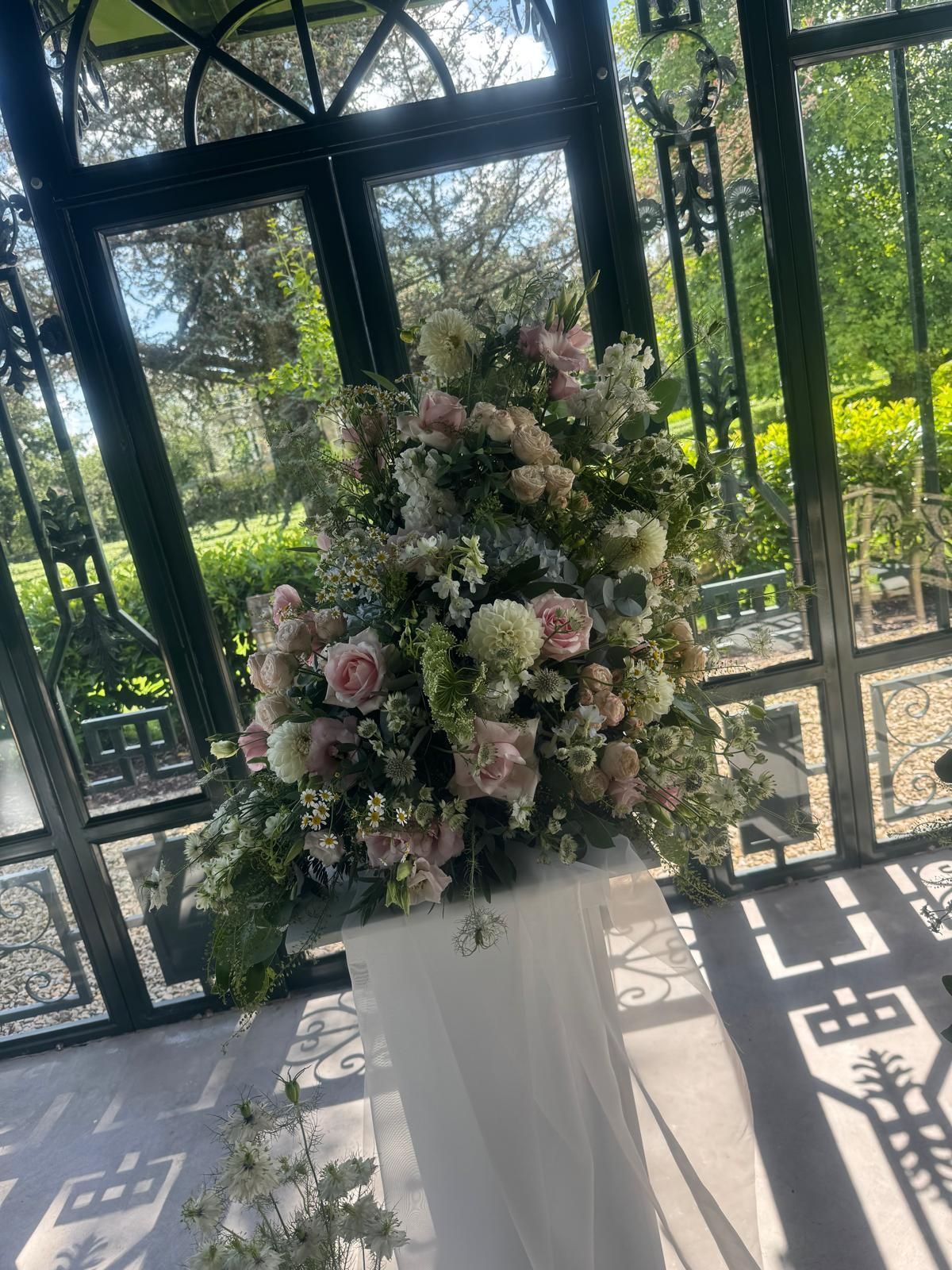 Large floral arrangement on a white pedestal, inside a glass-walled structure; pink and white flowers, greenery.