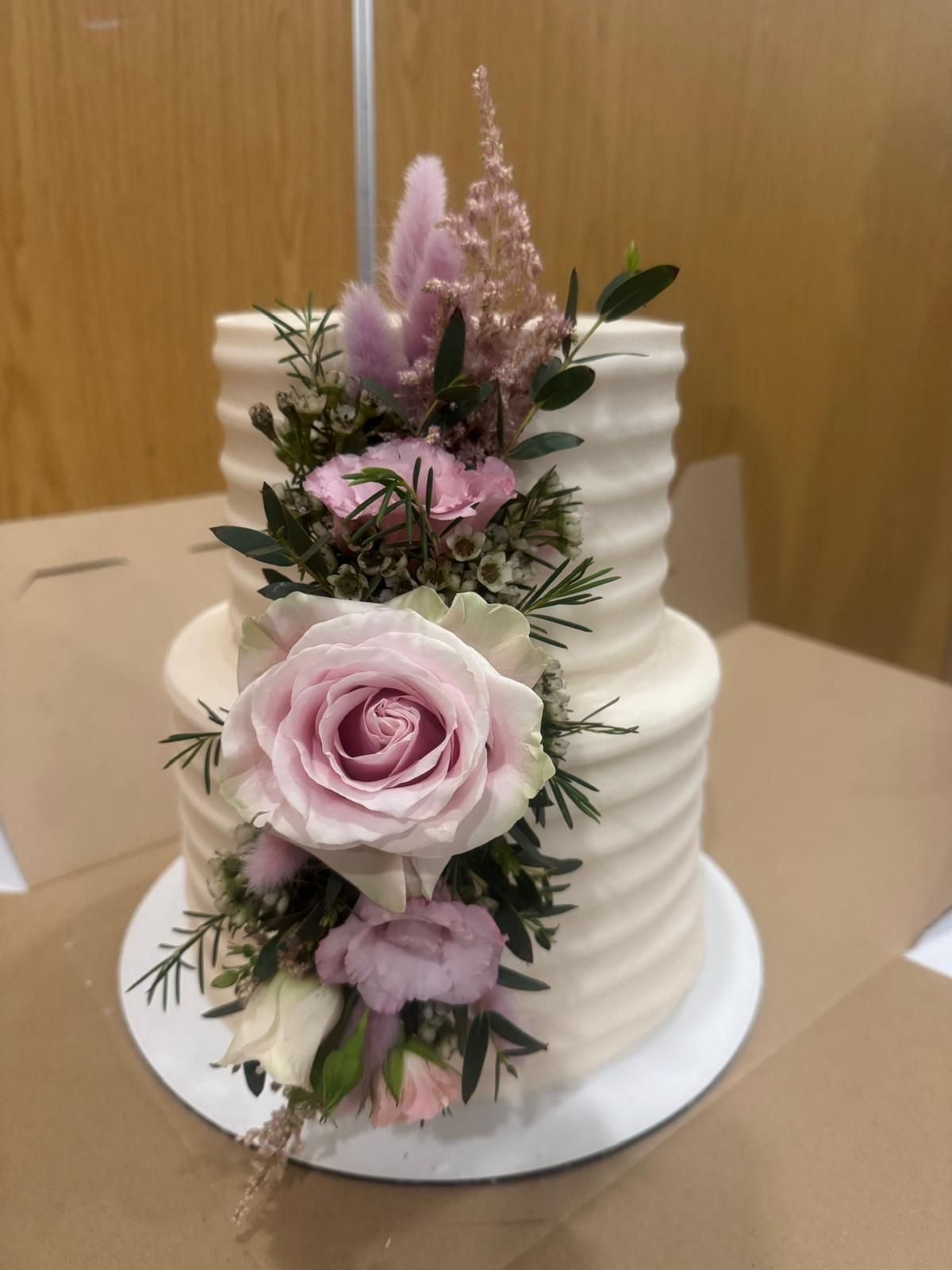 Two-tiered cake with cream frosting and floral arrangement of pink roses and greenery.