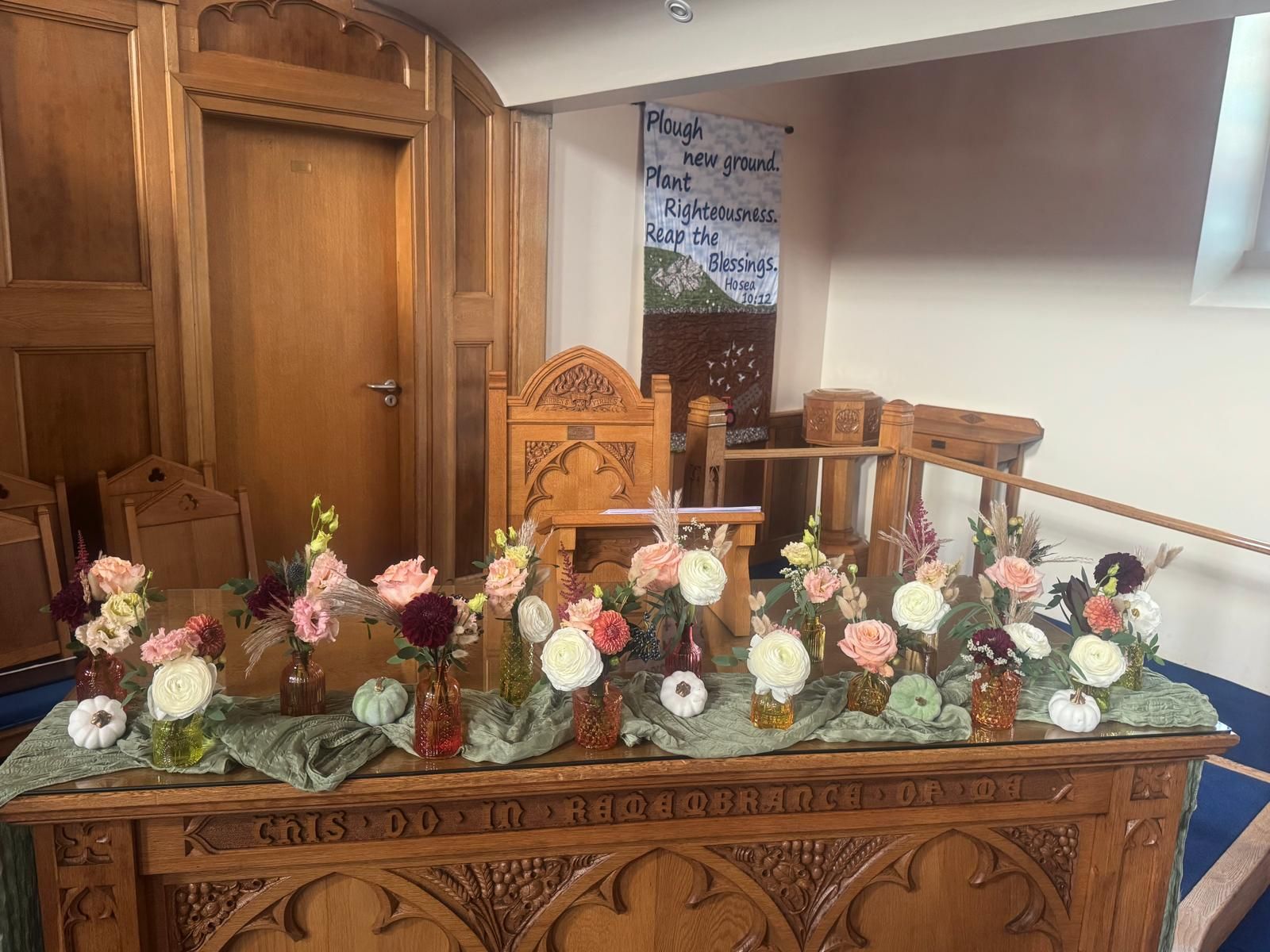 Altar adorned with flower arrangements; banner behind chair. Wooden furniture inside a church.