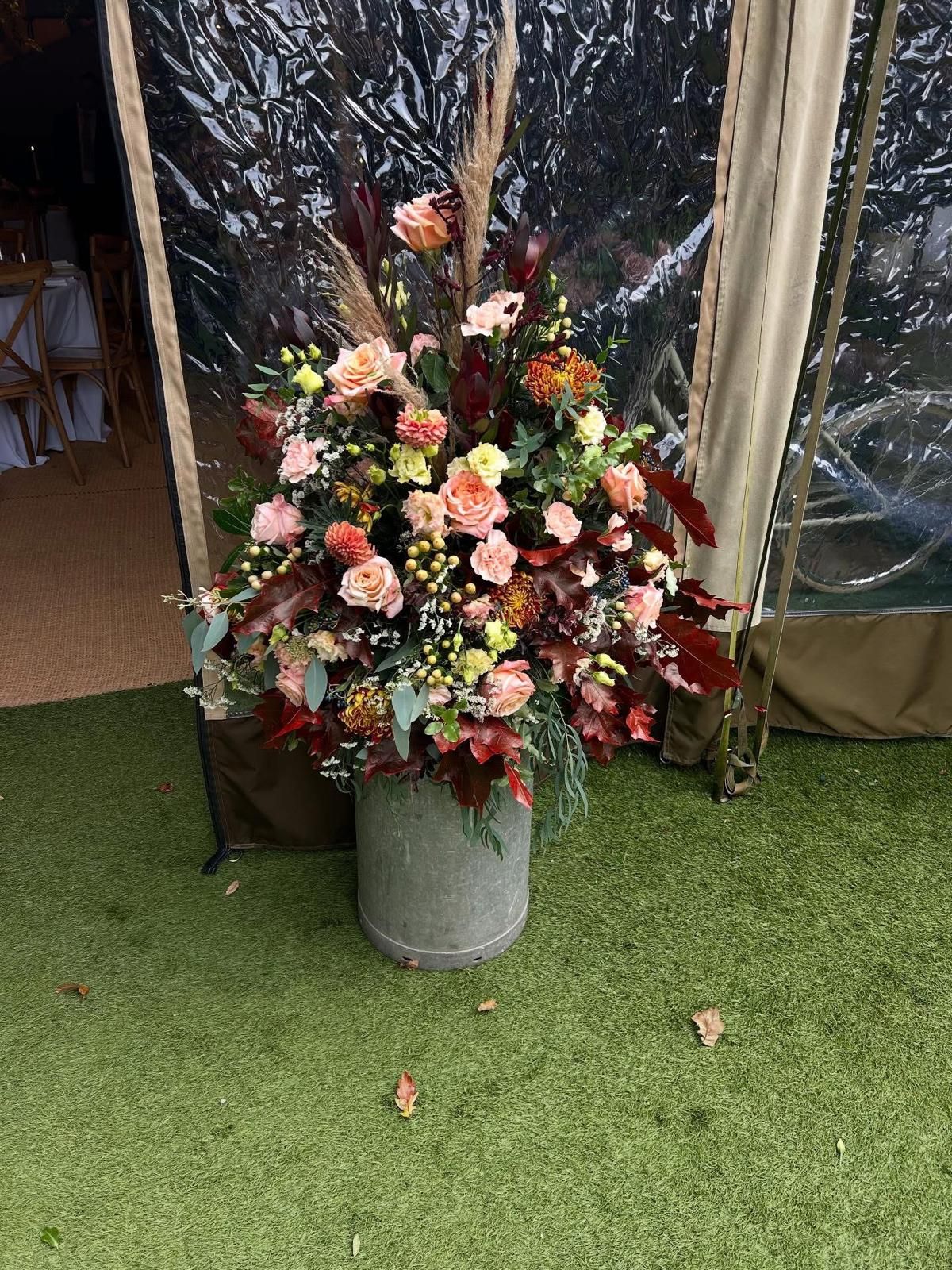 Large floral arrangement in a concrete pot, displayed on green turf next to a clear tent.