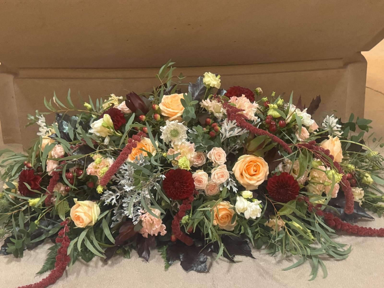 Floral arrangement with peach roses, red dahlias, and greenery on a beige surface.