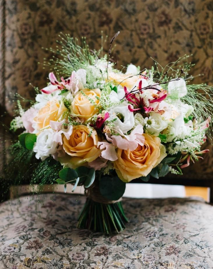 Bouquet of yellow and white roses, pink flowers, and greenery tied with twine, on a floral patterned chair.