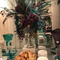 Elegant table centerpiece with tall purple-green floral arrangement, silver vases, teal decor, and pastries