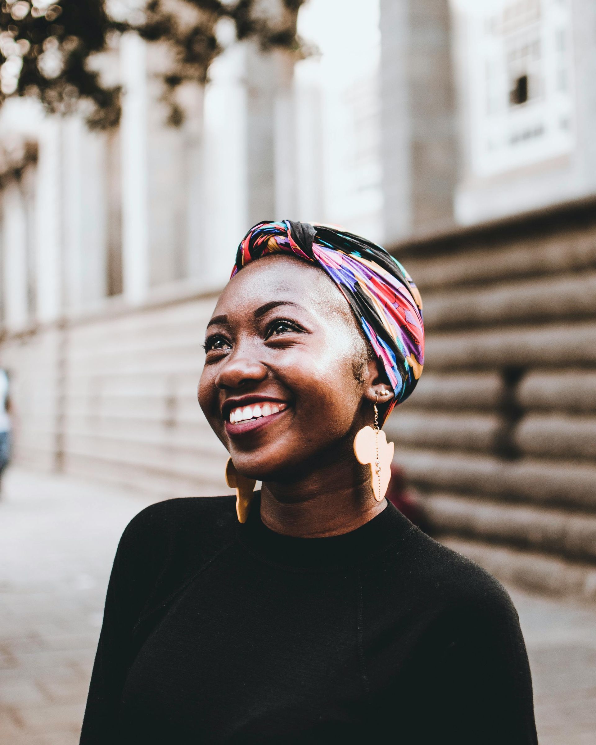A woman wearing a head scarf and earrings is smiling in front of a building.