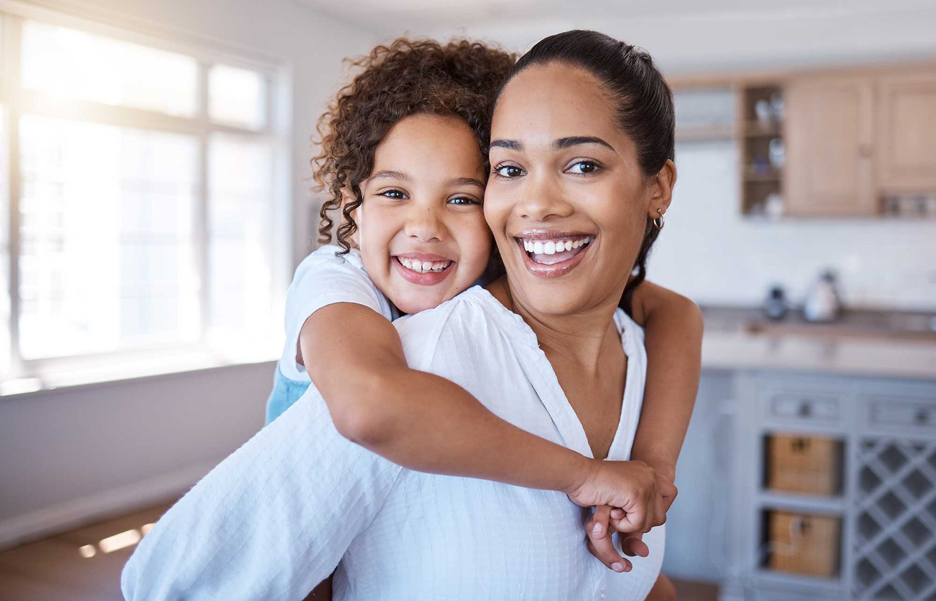 A woman is carrying a little girl on her back in a kitchen.