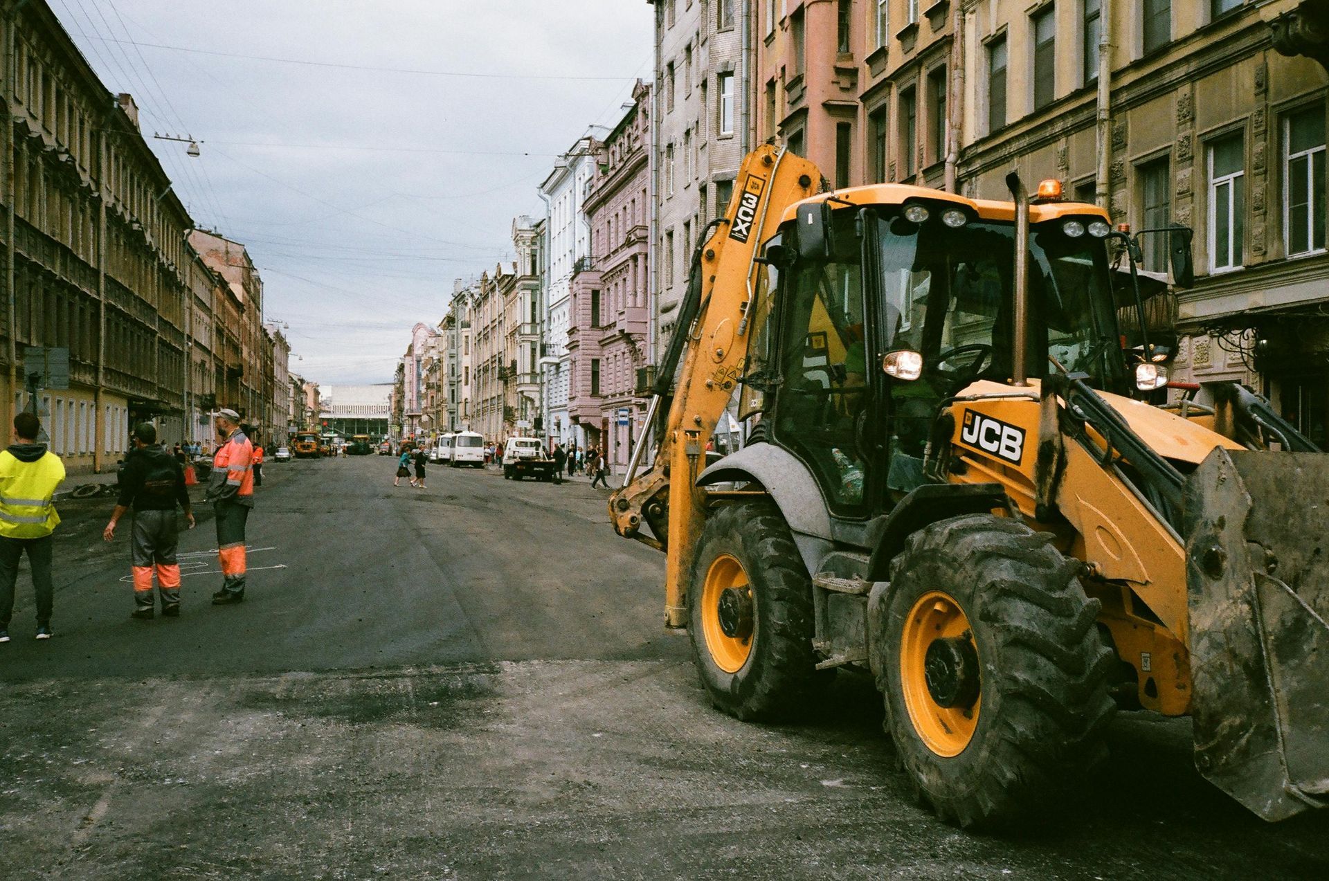 Yellow JCB backhoe on a city street; workers in safety vests; buildings in the background.