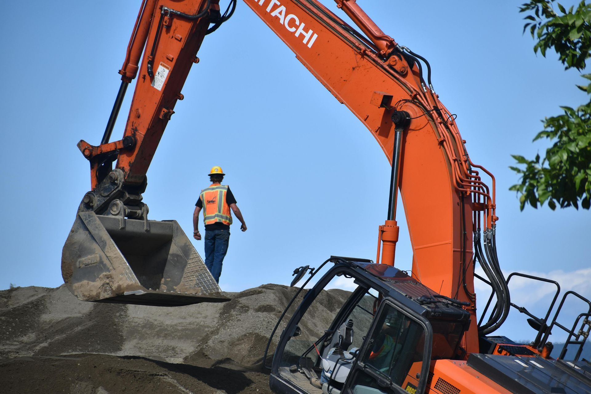 Orange Hitachi excavator with a worker in a hard hat, operating outdoors on a mound of dirt under a blue sky.